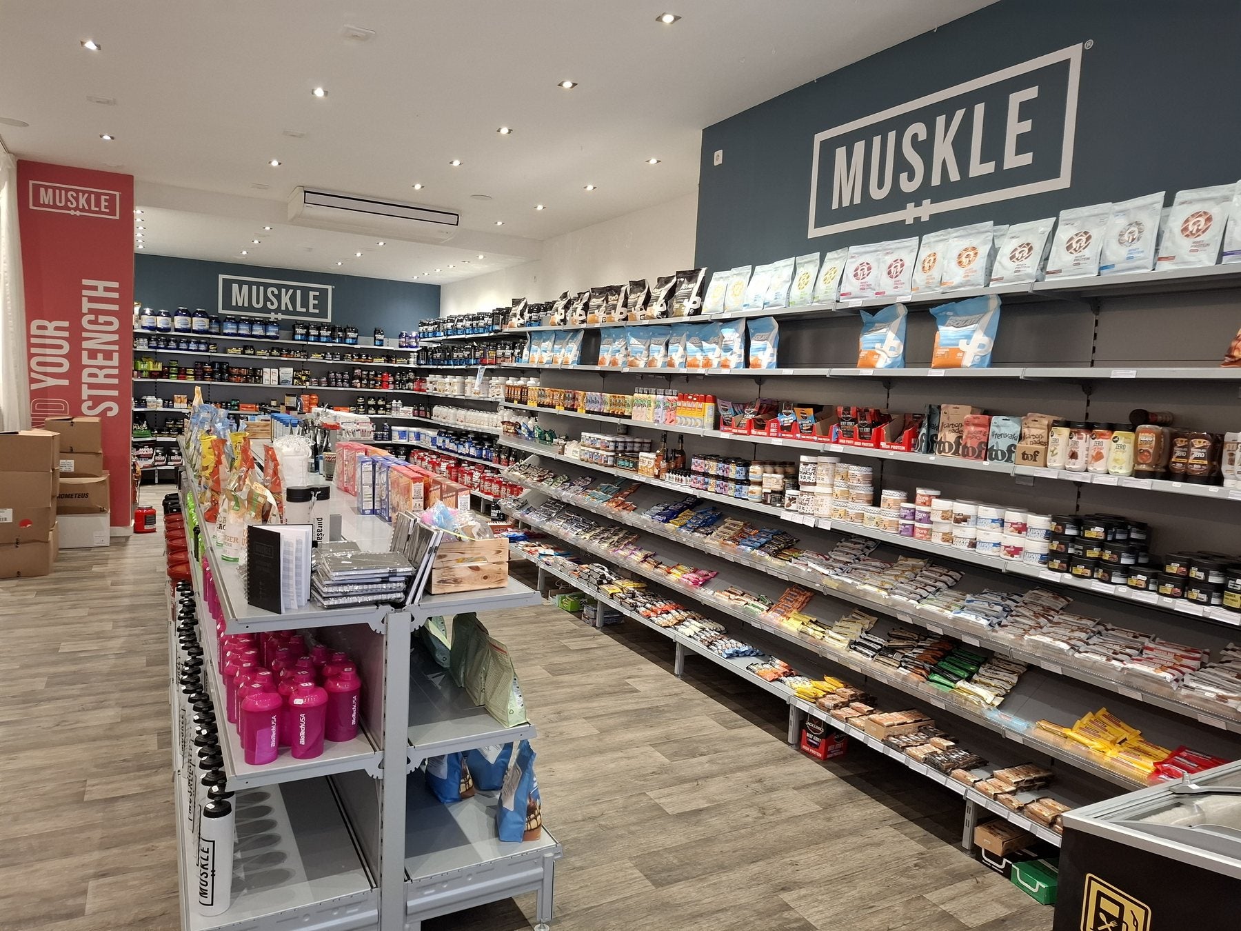 Winkelgang van MUSKLE Antwerpen met snackwand, shakers en rode BUILD YOUR STRENGTH-zuil