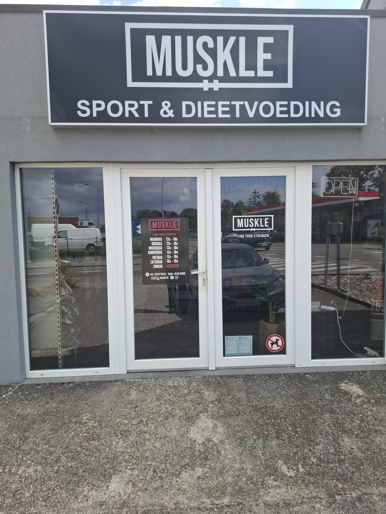 Ingang van MUSKLE Bonheiden met MUSKLE-fascia, openingsurenposter en OPEN-neon