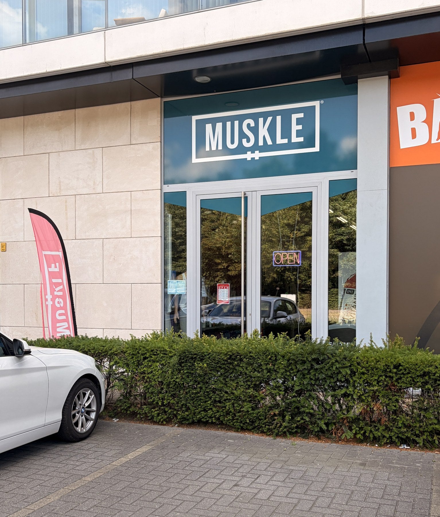 Ingang van MUSKLE Hasselt met teal MUSKLE-bord, glazen deuren en OPEN-bord